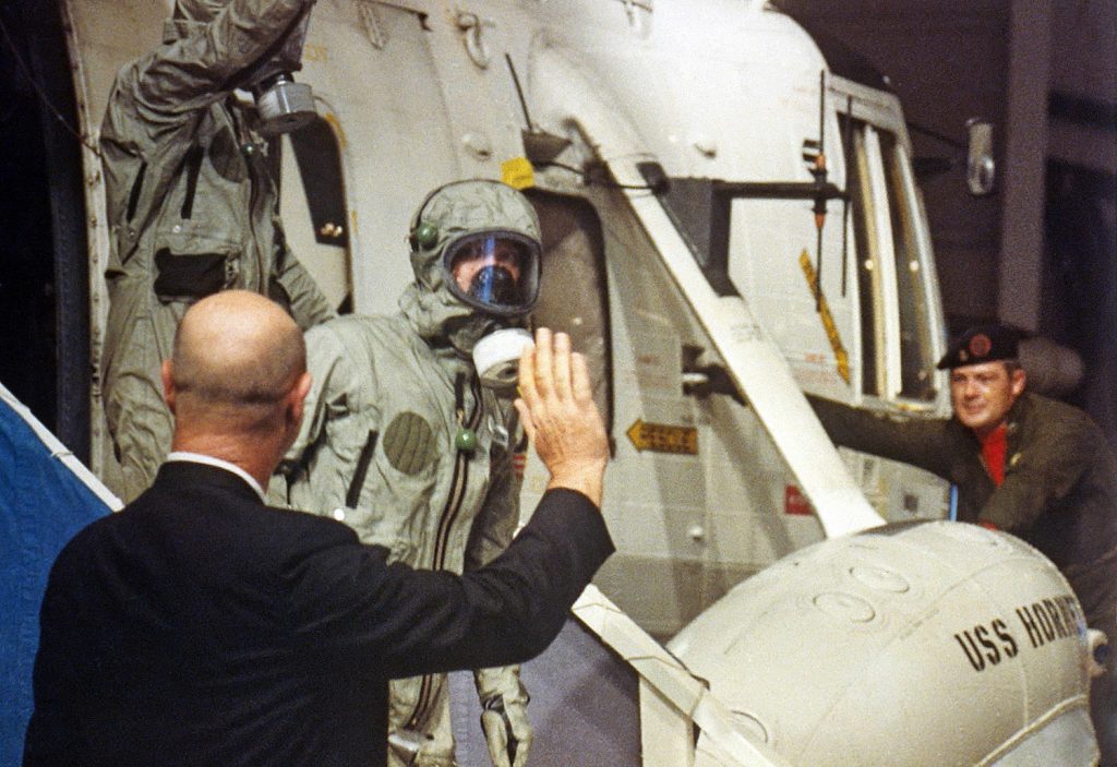 A member of the NASA recovery personnel waves to Apollo 11 crew (Michael Collins in front) as they depart the recovery helicopter aboard the USS Hornet.