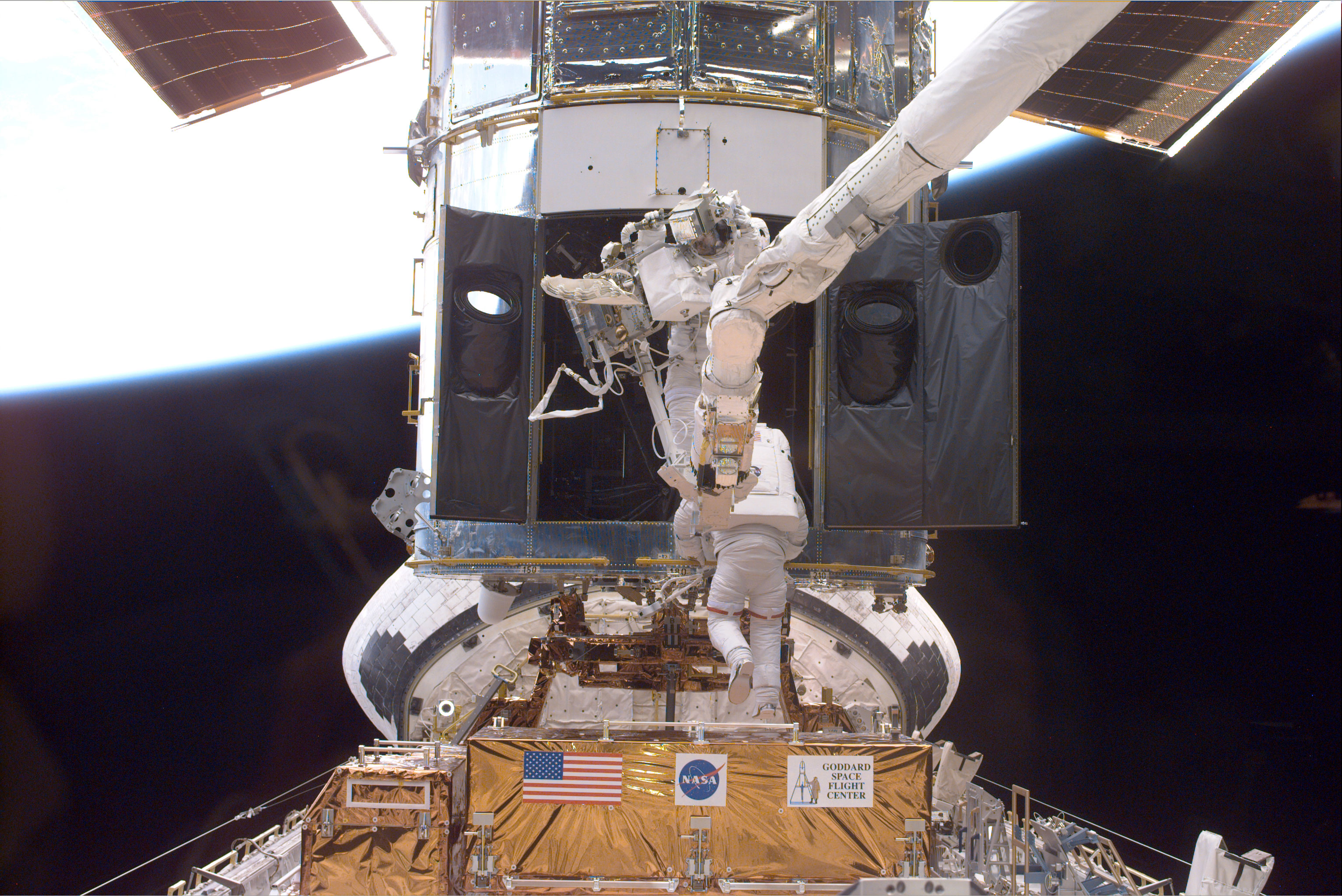 Payload Commander Steven L. Smith (bottom), and Mission Specialist John M. Grunsfeld, perform servicing tasks on the temporarily-captured Hubble Space Telescope. Grunsfeld is on a foot restraint connected to Discovery’s RMS robot arm. Smith, making his second servicing visit to HST, is using handrails on the telescope. Credit: NASA