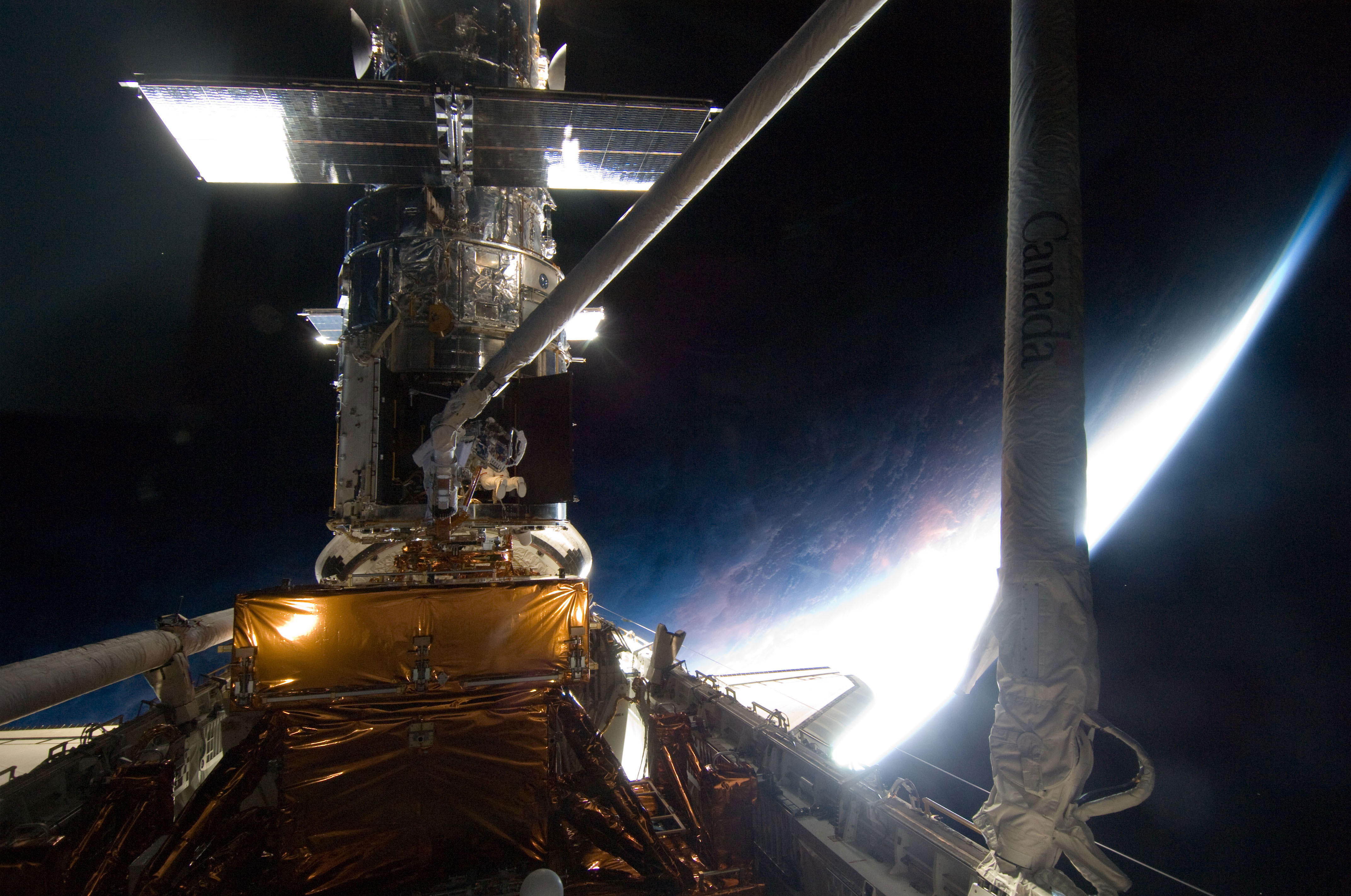 With a mostly dark home planet behind him, STS-125 mission specialist Michael Good rides Atlantis’ remote manipulator system arm to where he needs be while working on the Hubble Space Telescope. Credit: NASA