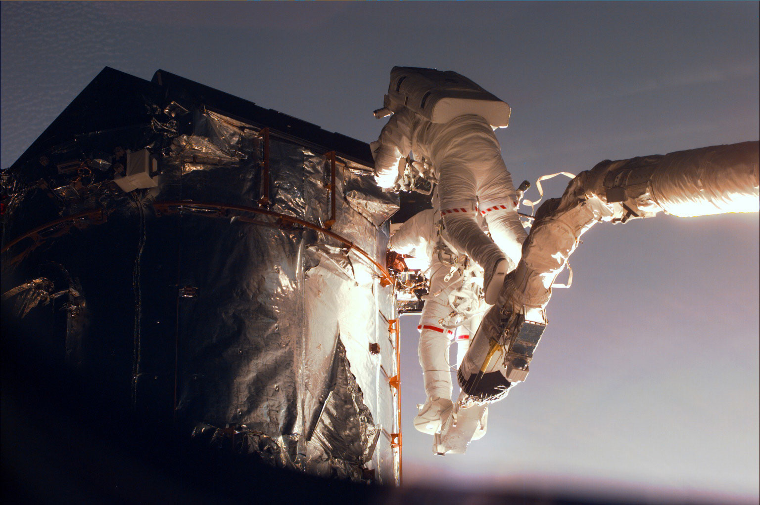 Gregory Harbaugh (solid stripe on EMU) uses the remote manipulator system (RMS) as a cherry-picker device to service Hubble. In cooperation with Joseph Tanner, nearby, Harbaugh is in the process of replacing the Magnetic Sensing System (MSS) protective caps with new, permanent covers. Credit: NASA