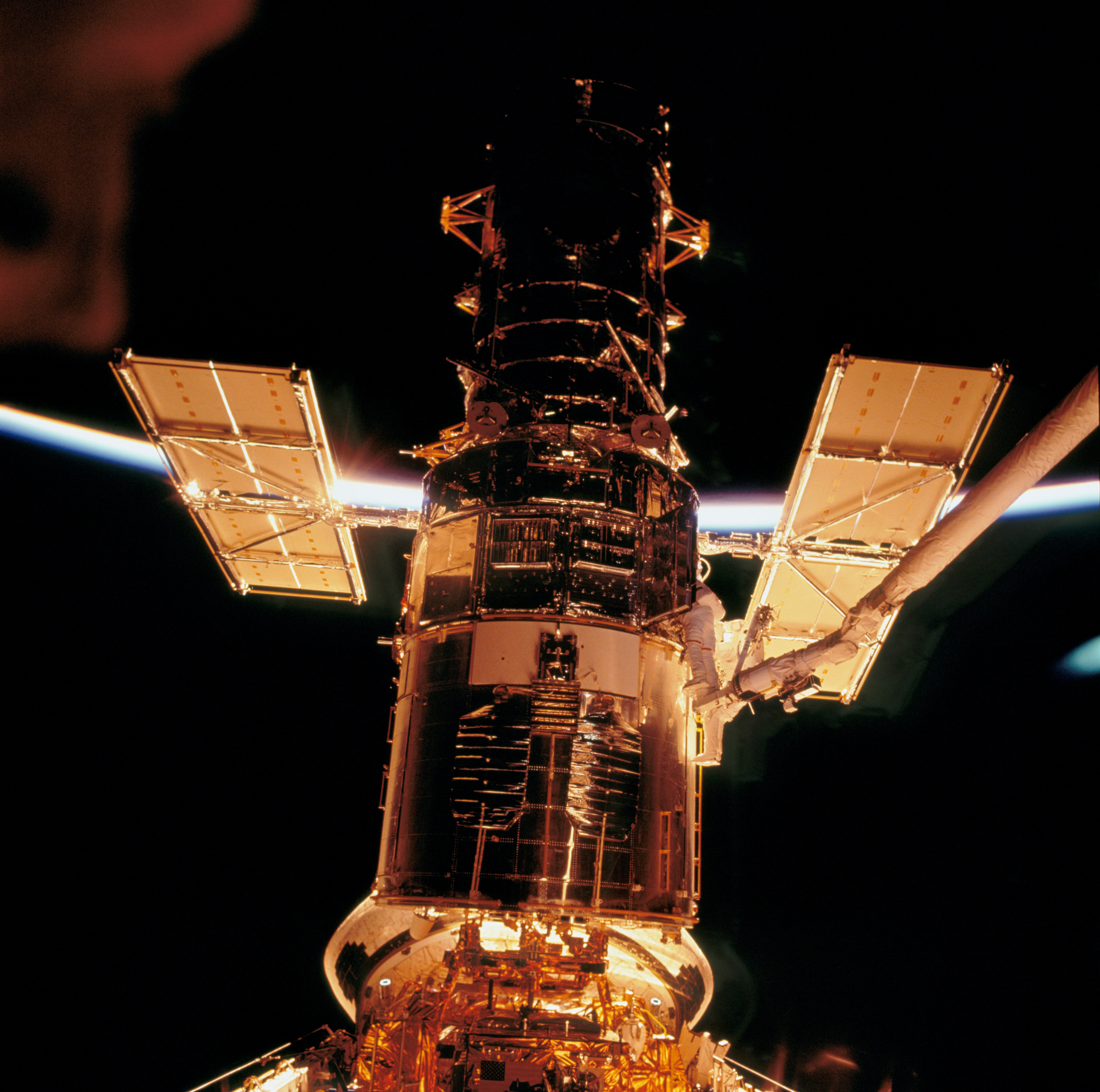 James Newman and Mike Massimino perform the first science instrument upgrade of the fourth Hubble Space Telescope servicing mission during the flight’s fourth EVA. Hubble, illuminated by the sunrise, provides stark contrast to the blackness of space in this photo. Arching between the telescope and one of the solar panels is the thin line of Earth’s atmosphere. Credit: NASA