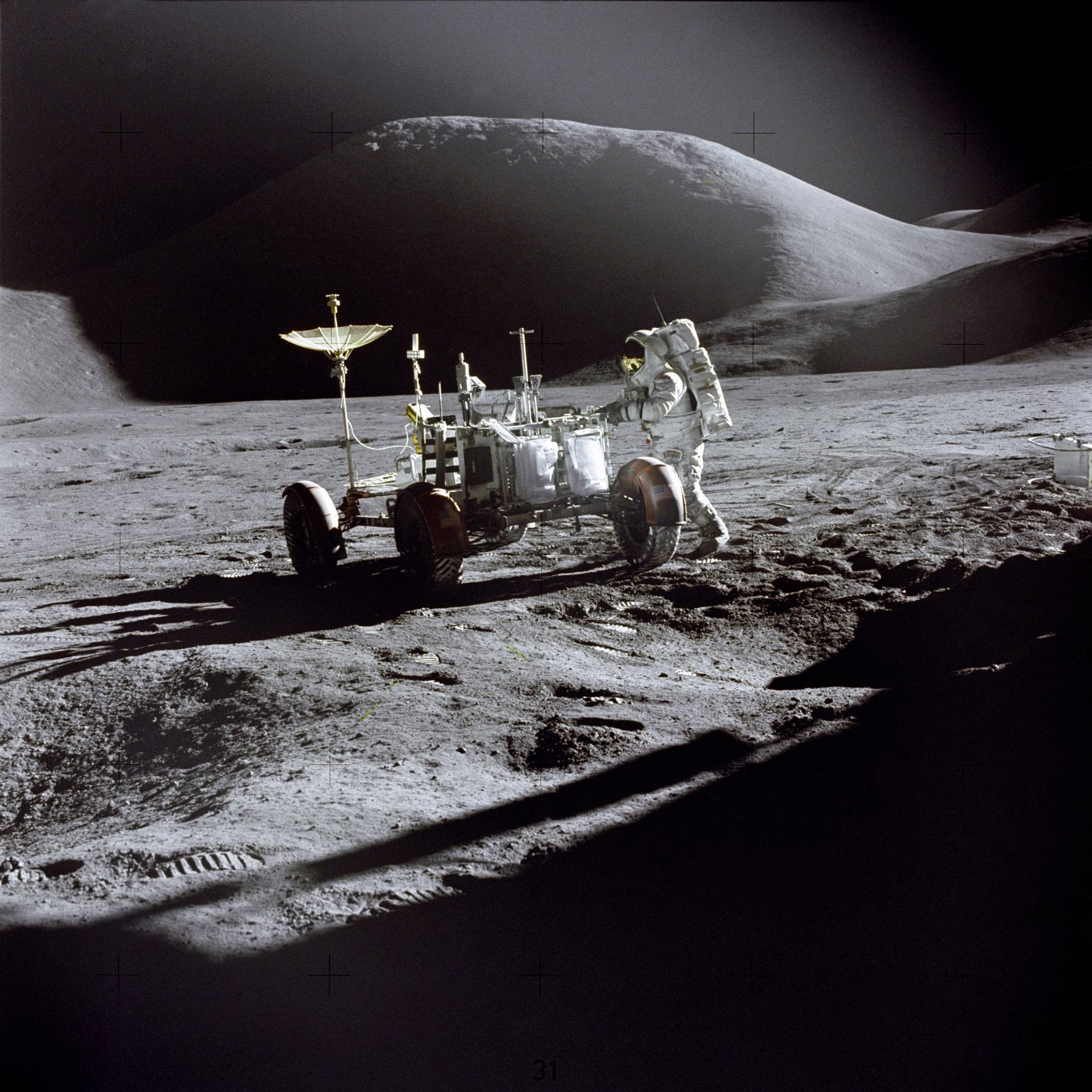 Jim Irwin is photographed beside the Lunar Roving Vehicle, with Mount Hadley in the background. Seen on the back of the Rover are two sample collection bags mounted on the gate, along with the rake, both pairs of tongs, the extension handle with scoop probably attached, and the penetrometer. Credit: NASA